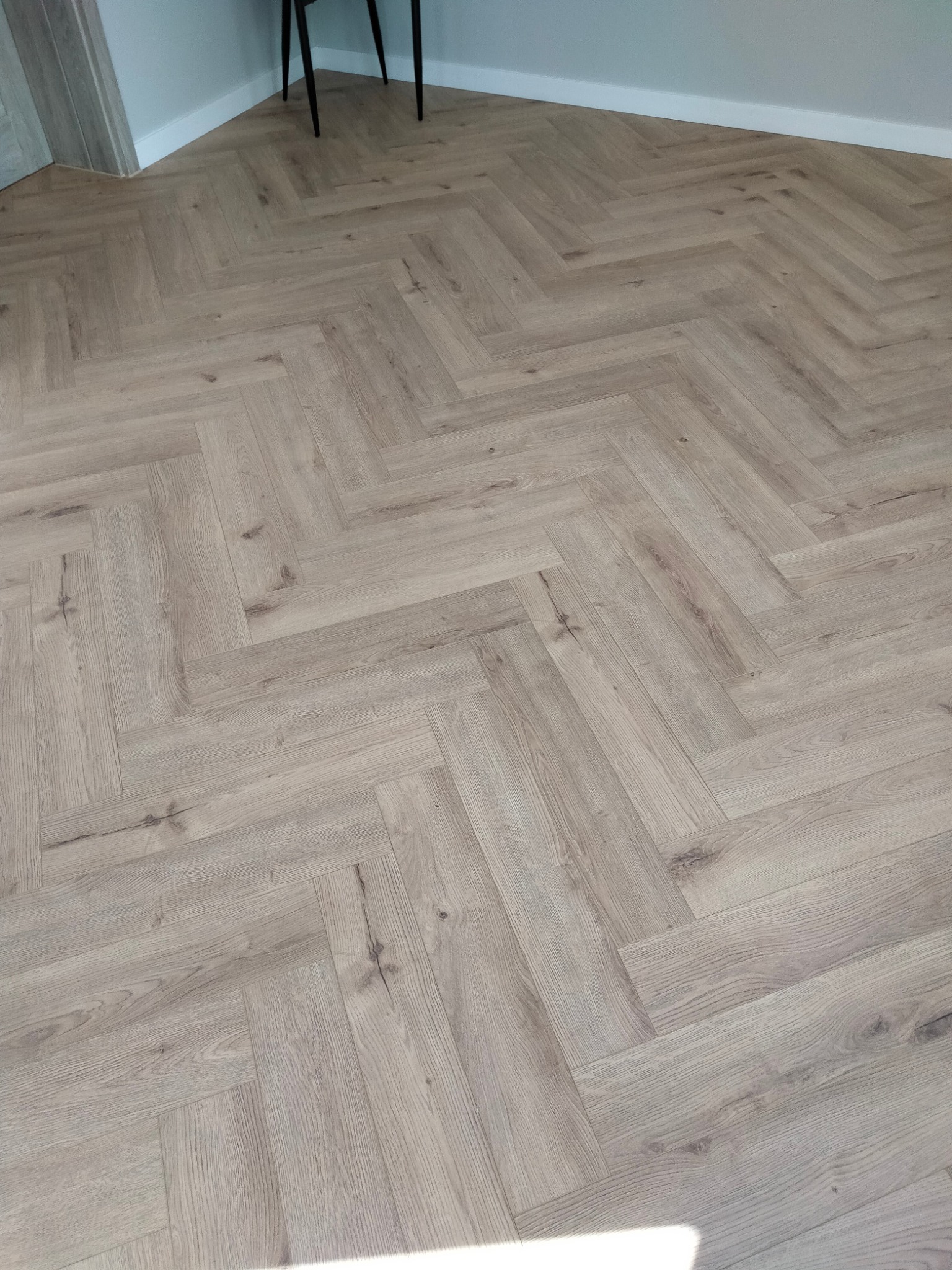 Jasne panele ułożone w jodełkę francuską, widoczne nogi krzesła i fragment listwy przypodłogowej. Naturalne światło podkreśla strukturę drewna.