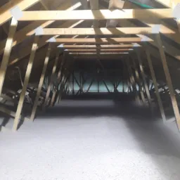Poddasze z drewnianą konstrukcją krokwiową wypełnione szarą izolacją celulozową, widoczne metalowe łączniki krokwi.