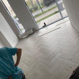 Łukasz Marchewka - Ułożona pod kątem jasna podłoga z płytek imitujących drewno w nowym mieszkaniu, widok na ulicę przez duże okno, fragment osoby w niebieskiej koszulce z plamami i plecak w rogu.