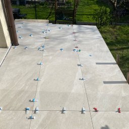 Łukasz Marchewka - Układanie beżowych płytek na tarasie z użyciem systemów poziomowania, widoczny fragment ogrodu w tle.