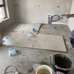 Łukasz Marchewka - Układanie płytek podłogowych w trakcie remontu, widoczne płytki z jasnego marmuru, niebieskie kliny poziomujące, wiadro z zaprawą, paca zębata i instalacja wodna w tle.