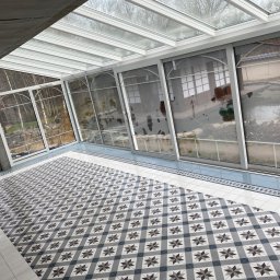 Łukasz Marchewka - Nowoczesna weranda z przeszklonym dachem i ścianami, wykończona dekoracyjną, wzorzystą podłogą z płytek w odcieniach szarości i bieli, widok na ogród z oczkiem wodnym.