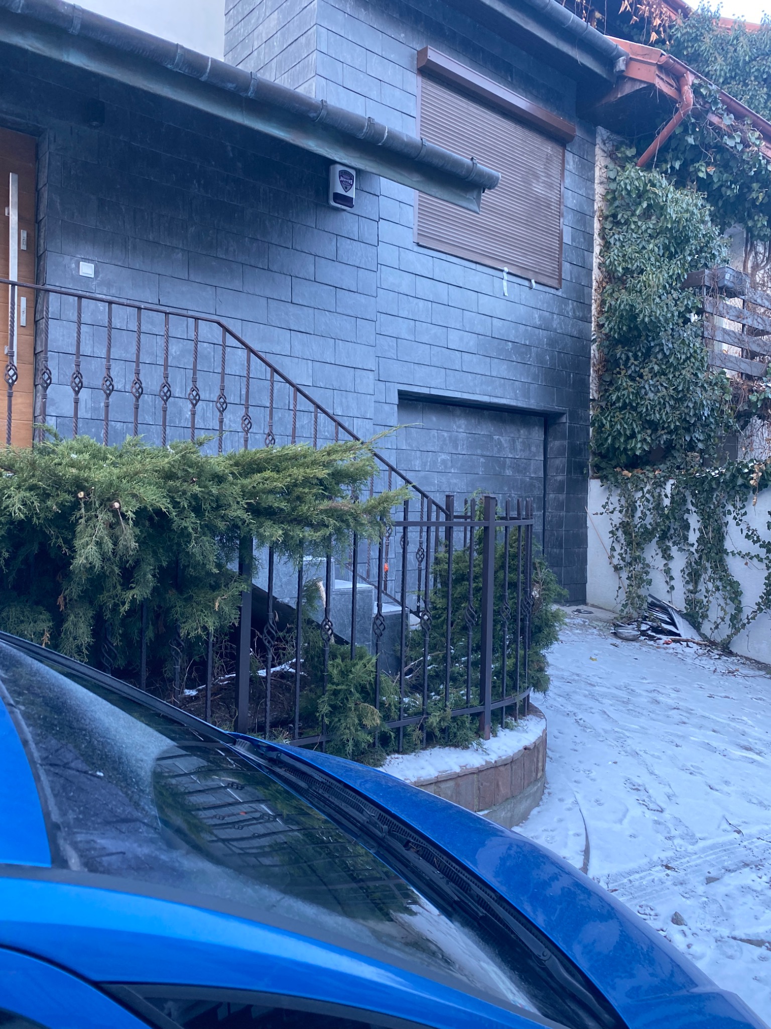 Dom z elewacją z ciemnego łupka, metalową balustradą schodów wejściowych i częściowo zasłoniętą roletą okienną, widziany zza niebieskiego samochodu w zimowej scenerii.