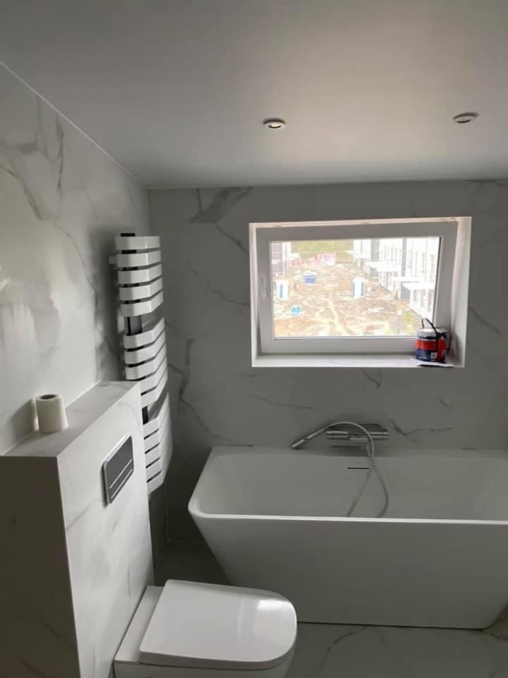 Nowoczesna łazienka wykończona w białym marmurze z wolnostojącą wanną, grzejnikiem drabinkowym i podwieszaną toaletą; za oknem widok na budowę.