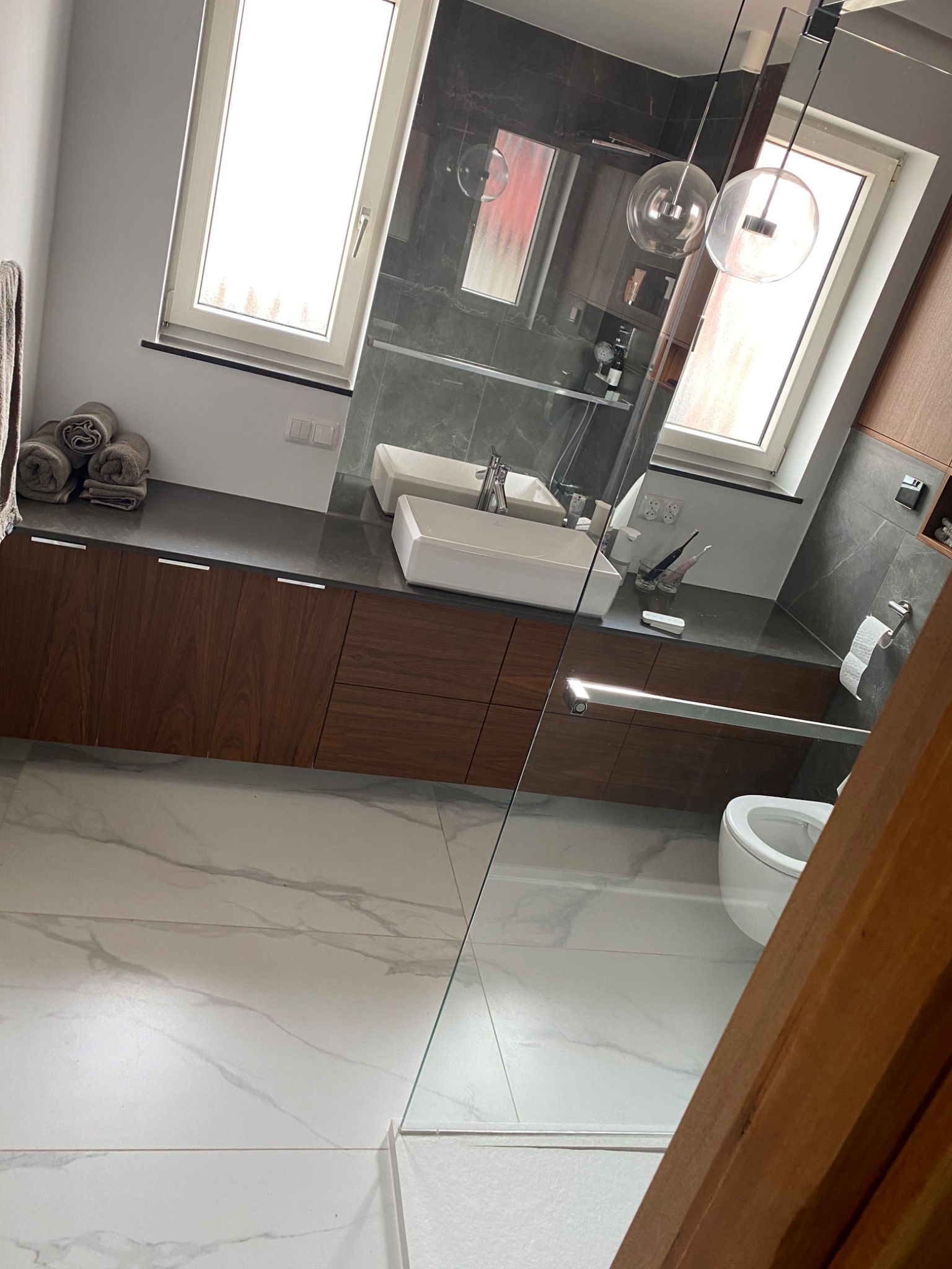 Nowoczesna łazienka z marmurową podłogą, drewnianymi szafkami pod umywalką, szklanym panelem prysznicowym i okrągłymi lampami wiszącymi.