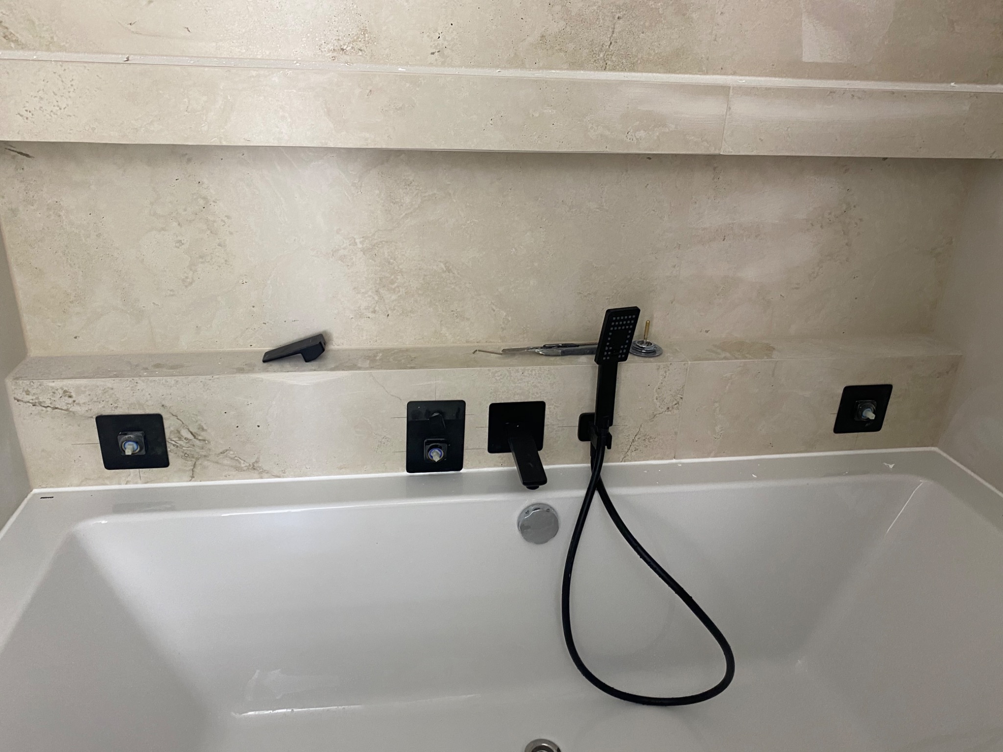 Nowoczesna biała wanna z czarną armaturą zamontowaną w beżowej, marmurkowej wnęce ściennej z półką i narzędziami