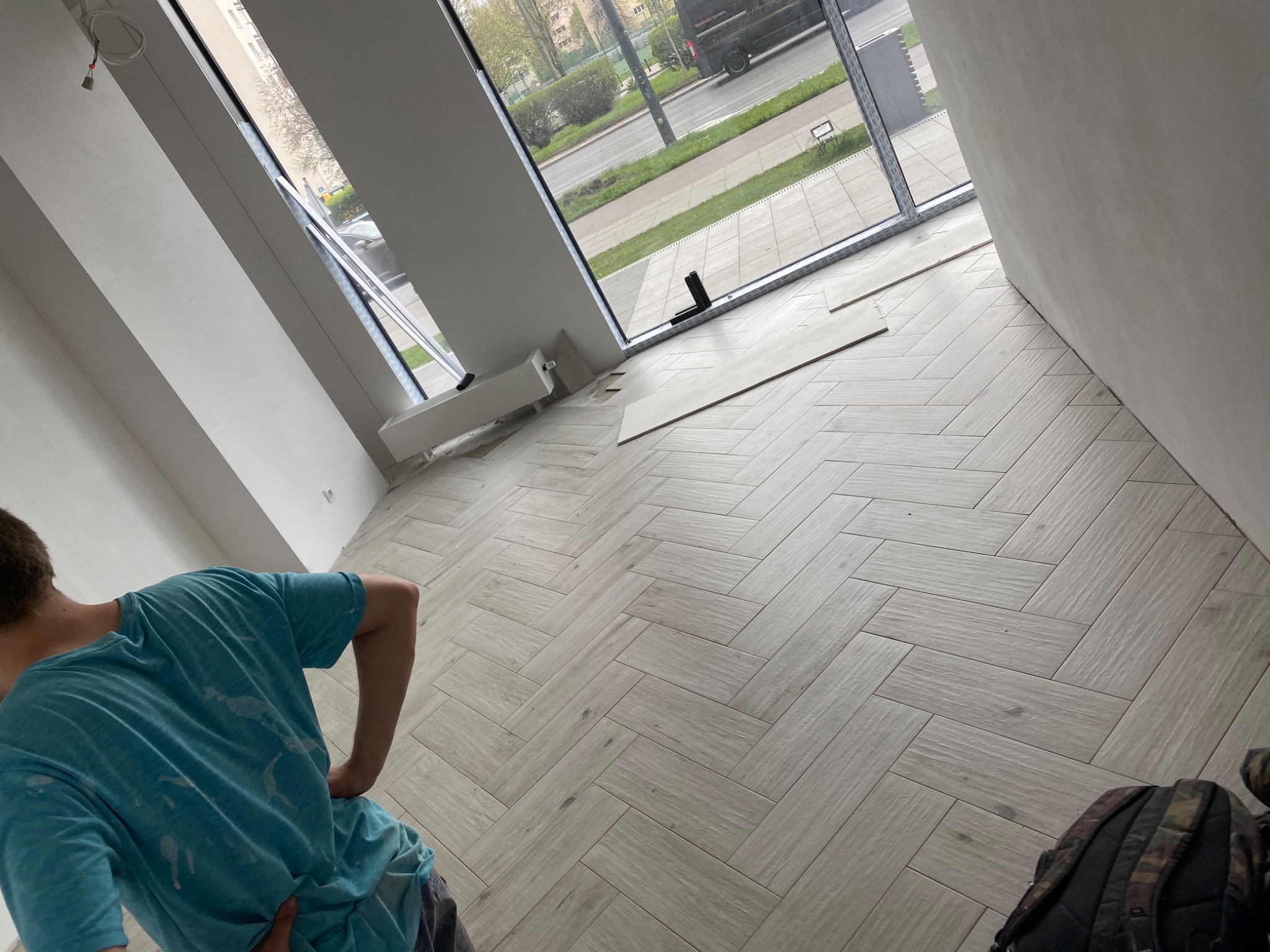 Ułożona pod kątem jasna podłoga z płytek imitujących drewno w nowym mieszkaniu, widok na ulicę przez duże okno, fragment osoby w niebieskiej koszulce z plamami i plecak w rogu.