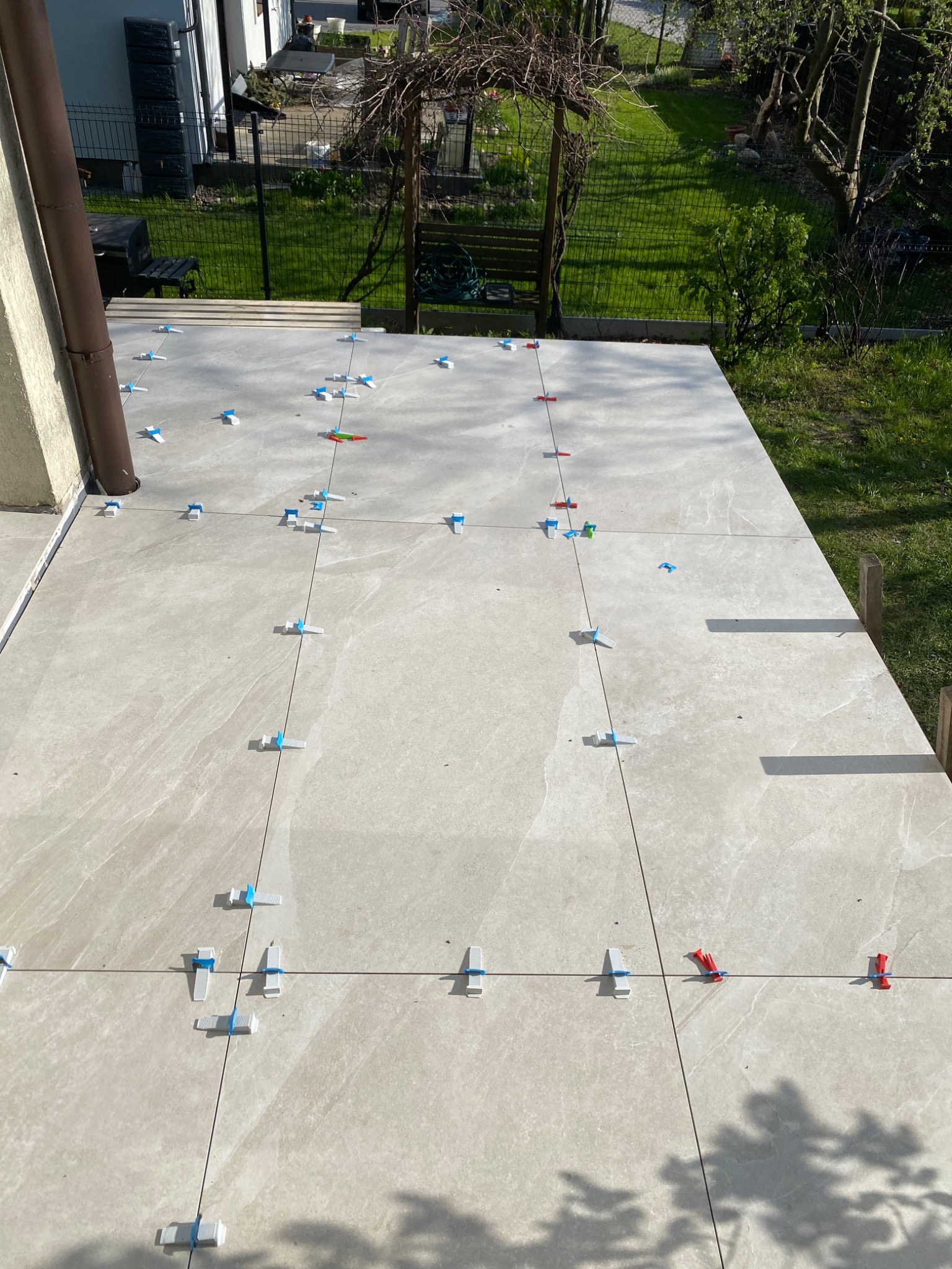 Układanie beżowych płytek na tarasie z użyciem systemów poziomowania, widoczny fragment ogrodu w tle.