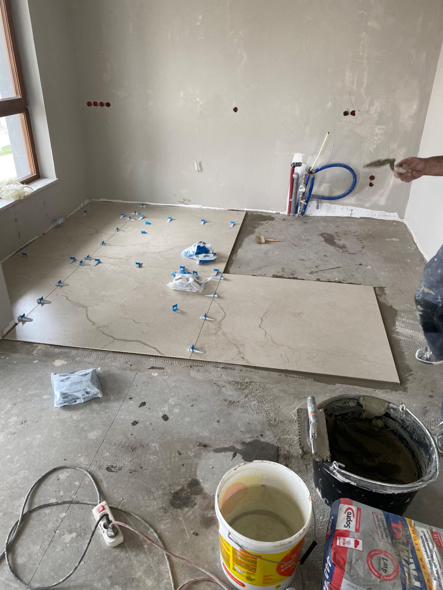Układanie płytek podłogowych w trakcie remontu, widoczne płytki z jasnego marmuru, niebieskie kliny poziomujące, wiadro z zaprawą, paca zębata i instalacja wodna w tle.