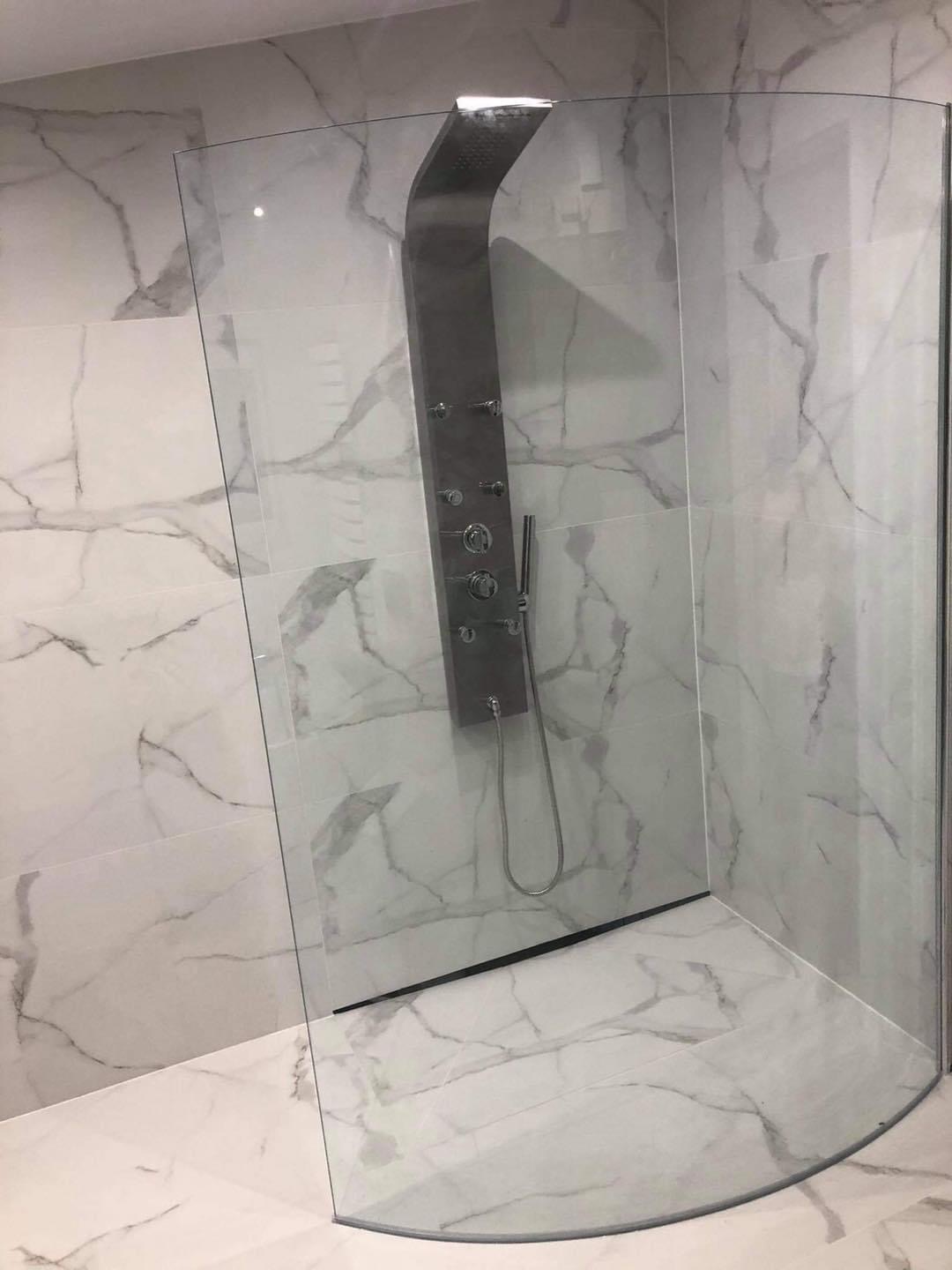 Nowoczesna kabina prysznicowa z panelem natryskowym i zakrzywioną szybą, wykończona białymi płytkami imitującymi marmur.