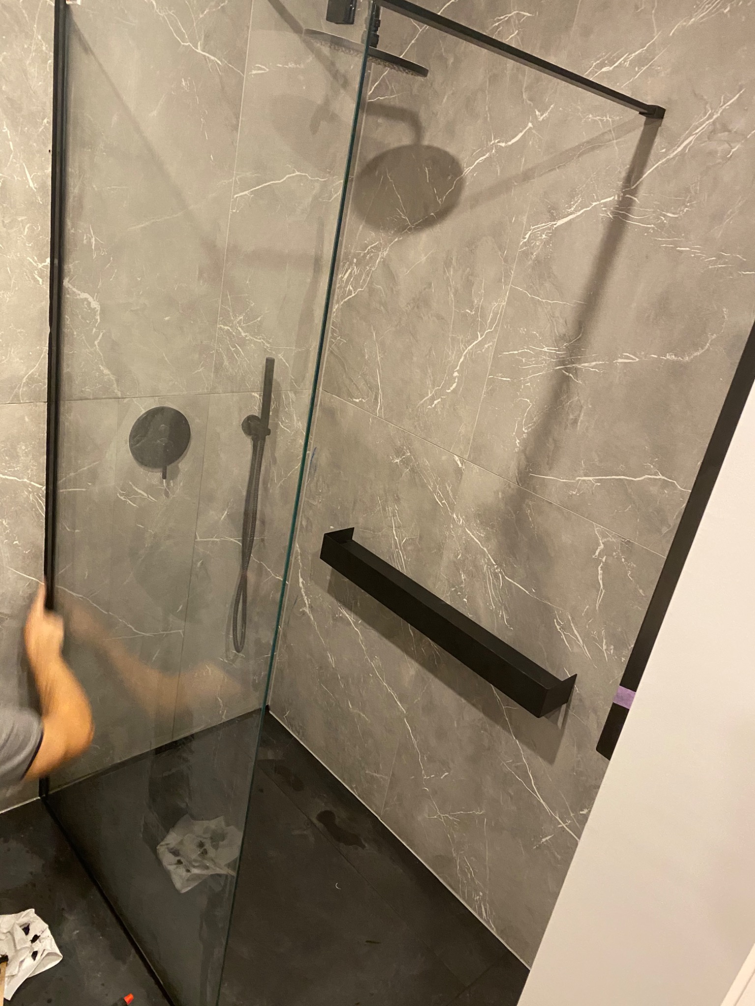 Montaż kabiny prysznicowej typu walk-in z czarnymi profilami na tle szarych płytek imitujących marmur, widoczne czarne elementy armatury.