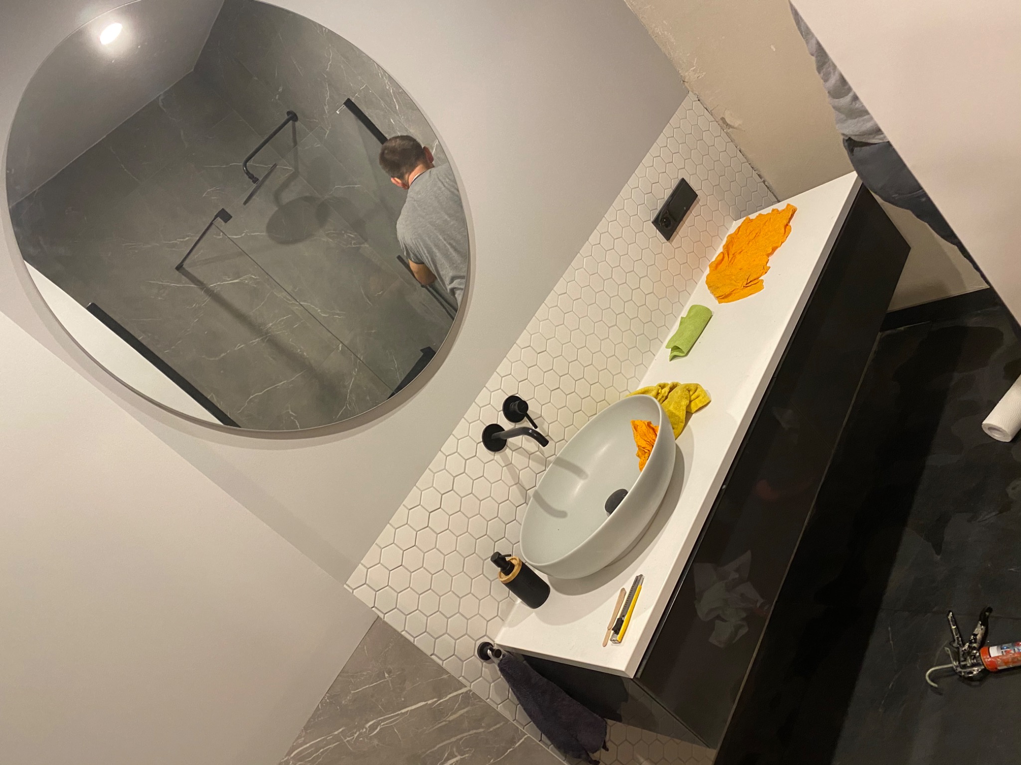 Łazienka w trakcie wykańczania: okrągłe lustro odbija kabinę prysznicową z szarymi płytkami imitującymi marmur, biała umywalka na blacie z czarnymi szafkami, białe heksagonalne płytki na ścianie...