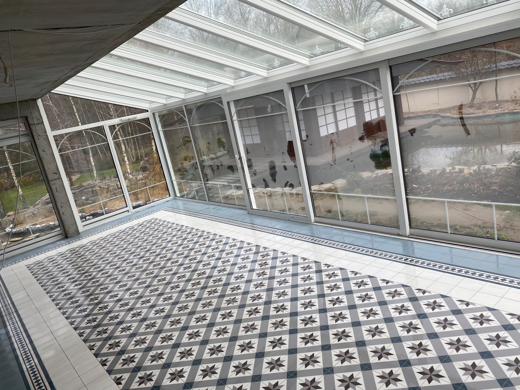 Nowoczesna weranda z przeszklonym dachem i ścianami, wykończona dekoracyjną, wzorzystą podłogą z płytek w odcieniach szarości i bieli, widok na ogród z oczkiem wodnym.