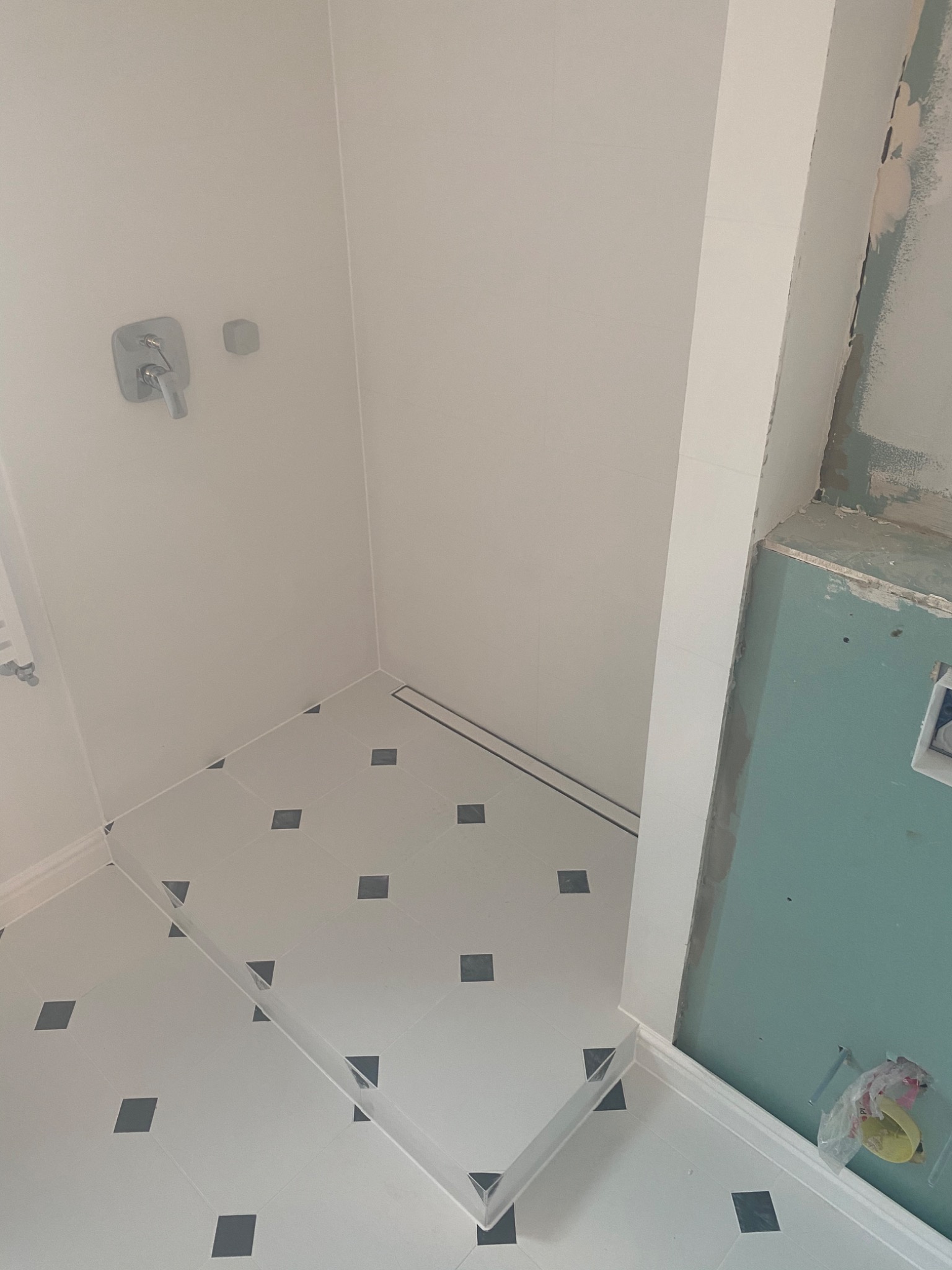 Narożnik kabiny prysznicowej z wyłożonymi białymi płytkami z ciemnymi kwadratami, widoczny odpływ liniowy i bateria, ściana w trakcie wykańczania z zieloną płytą gipsową.