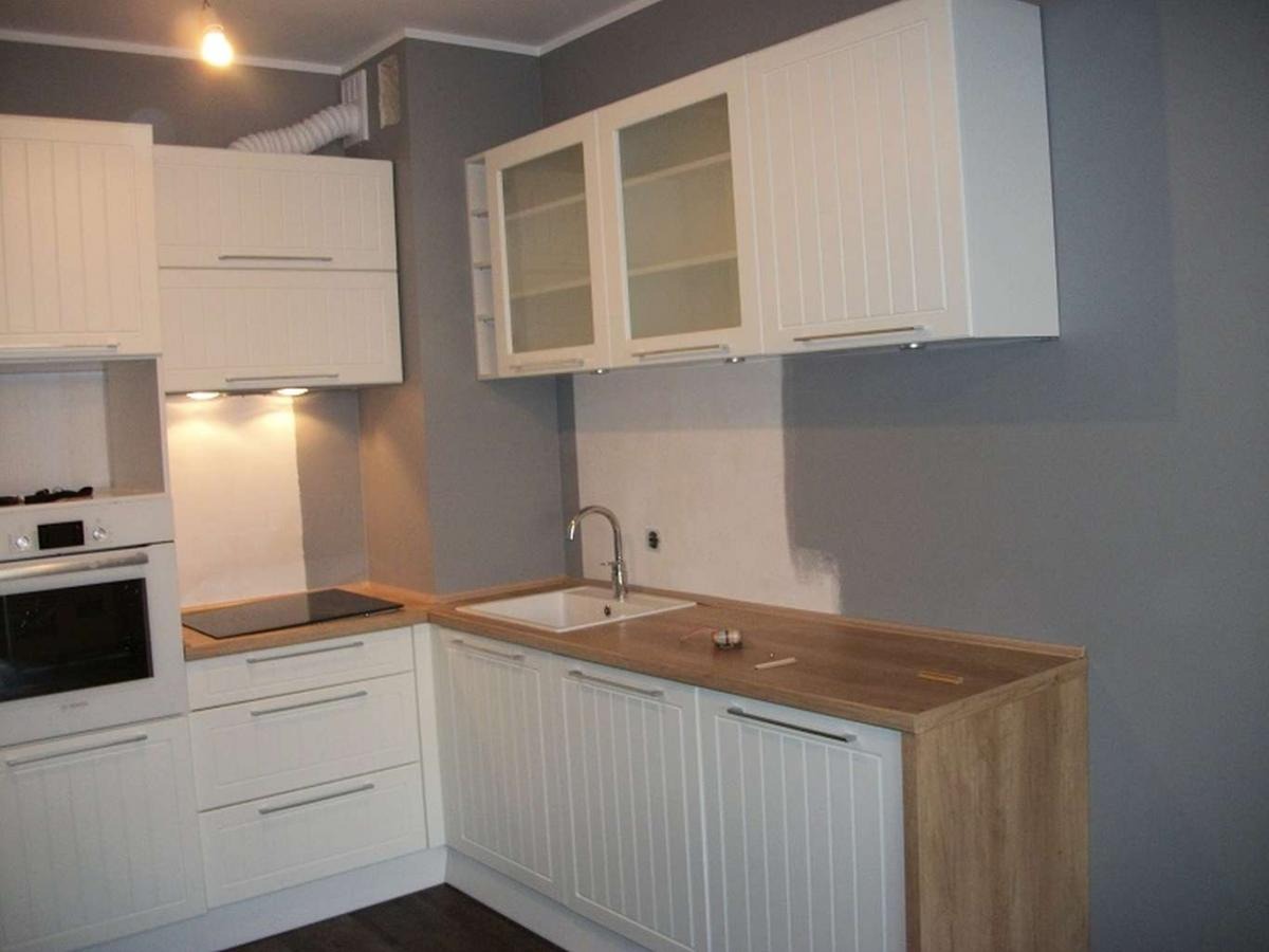 Jasna kuchnia z białymi frontami szafek z frezowanymi pionowymi liniami i drewnianym blatem; widoczny fragment ściany pomalowanej na szaro, biały okap z podłączonym elastycznym przewodem...