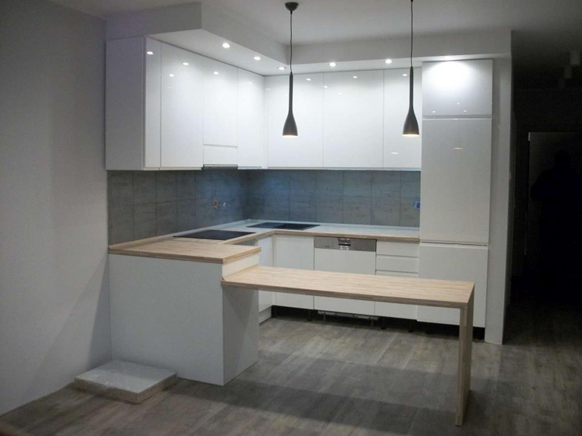 Nowoczesna, biała kuchnia z połyskiem, wyposażona w blat z jasnego drewna i minimalistyczne lampy wiszące, z płytkami w odcieniach szarości nad blatem roboczym.