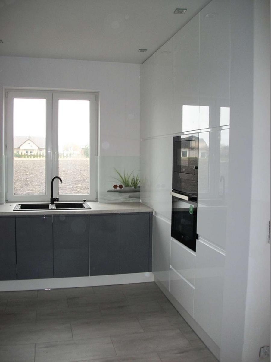 Nowoczesna kuchnia z szarymi szafkami dolnymi i białymi szafkami górnymi na wysoki połysk, czarny zlew i bateria, z oknem z widokiem na dom i pole.