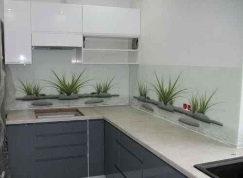 Nowoczesna kuchnia narożna z szarymi frontami i białymi szafkami wiszącymi, wyróżniająca się szklanym panelem nad blatem z motywem roślin i kamieni, imitującym krajobraz wodny.