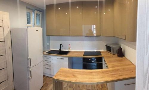 Nowoczesna kuchnia z beżowymi, lakierowanymi szafkami górnymi, białymi szafkami dolnymi i drewnianym blatem, czarnym zlewem i płytą grzewczą, oraz białą lodówką.