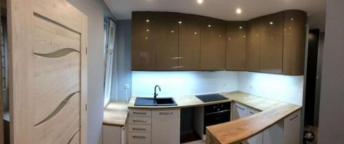 Nowoczesna kuchnia na wymiar z błyszczącymi, brązowymi szafkami górnymi i jasnymi szafkami dolnymi, czarny zlew i płyta indukcyjna, drewniany blat oraz drzwi wewnętrzne z jasnego drewna...