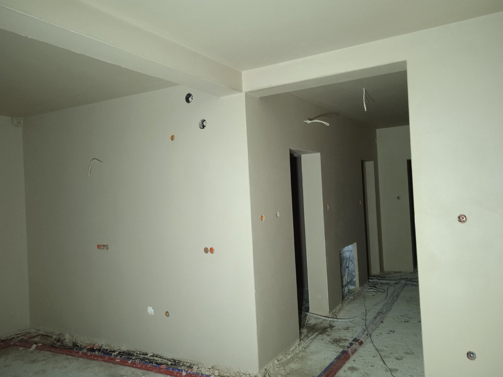 Wnętrze w trakcie prac wykończeniowych, ściany pokryte jasnym tynkiem, widoczne puszki elektryczne i wystające przewody, podłoga z resztkami zaprawy i przewodami instalacji.