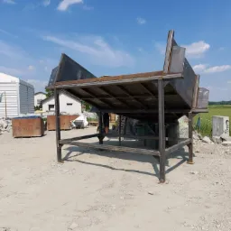 Konstrukcja metalowa z blachy, prawdopodobnie element bramy przemysłowej, w surowym stanie, na placu budowy z widocznymi blokami kamienia i budynkami w tle.