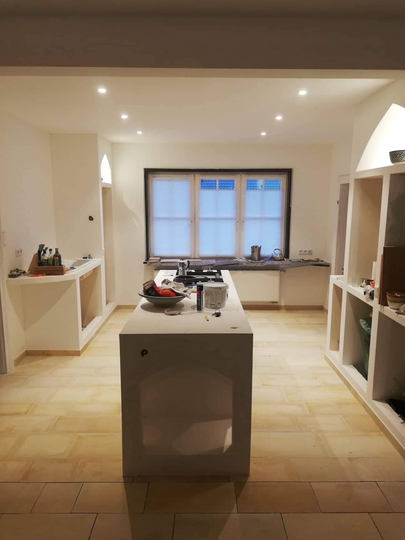 Wnętrze kuchni w trakcie prac wykończeniowych, z wyspą na środku, zabudowanymi wnękami po bokach i oknem z roletami w tle, widoczne narzędzia i materiały malarskie na wyspie, ściany przygotowane...