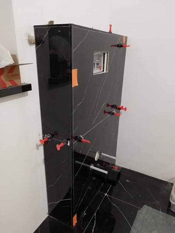 Montaż stelaża podtynkowego WC z czarnymi płytkami imitującymi marmur, z użyciem poziomowania krzyżykami i klinami, widoczne rury instalacyjne.