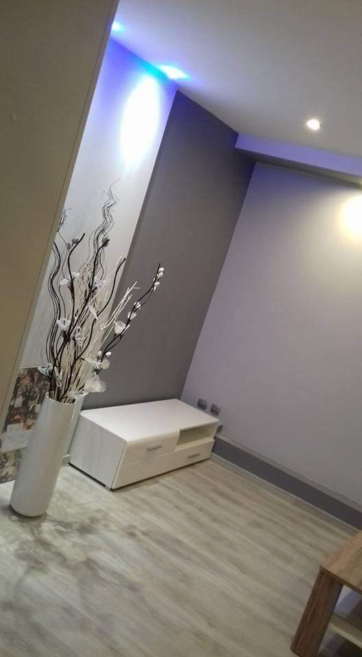 Nowoczesne wnętrze z szarą ścianą, białym meblem RTV i wazonem z dekoracyjnymi gałązkami, oświetlone niebieskim światłem LED na suficie.