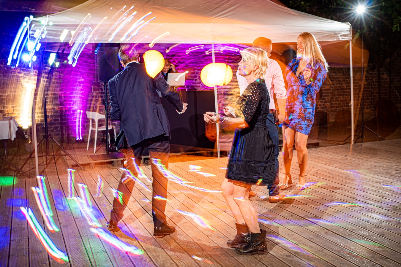 Goście weselni tańczą na parkiecie pod namiotem, oświetlonym kolorowymi reflektorami, w tle DJ i ceglana ściana.