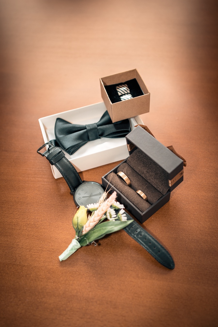 Przygotowania do ślubu: złote obrączki w pudełku, czarna mucha, spinki do mankietów w pudełku, zegarek z butonierką na drewnianym stole.