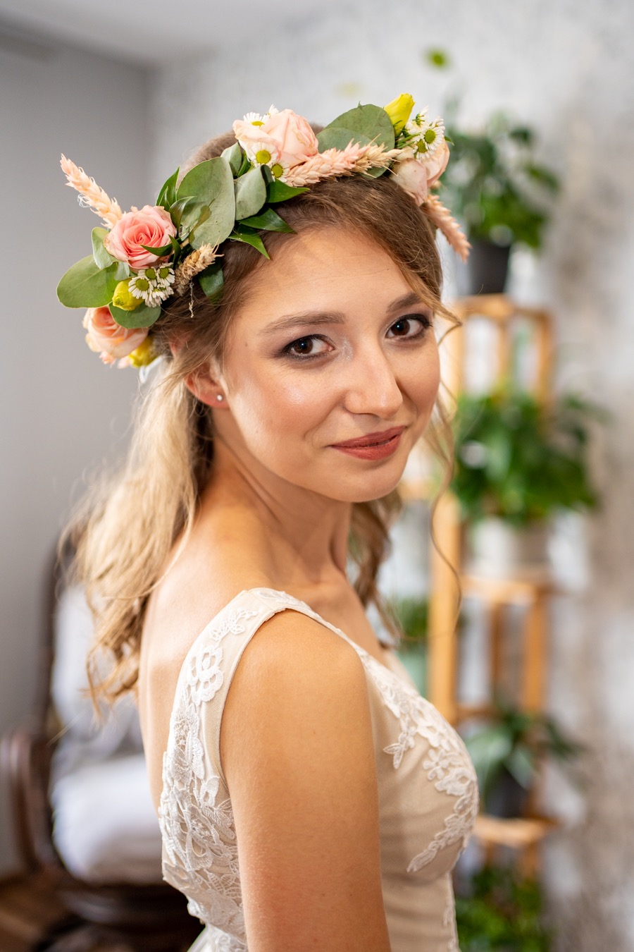 Portret uśmiechniętej kobiety w białej sukni z koronkowymi detalami i wiankiem z różowych i białych kwiatów na głowie, delikatny makijaż, w tle drewniany regał z roślinami doniczkowymi.