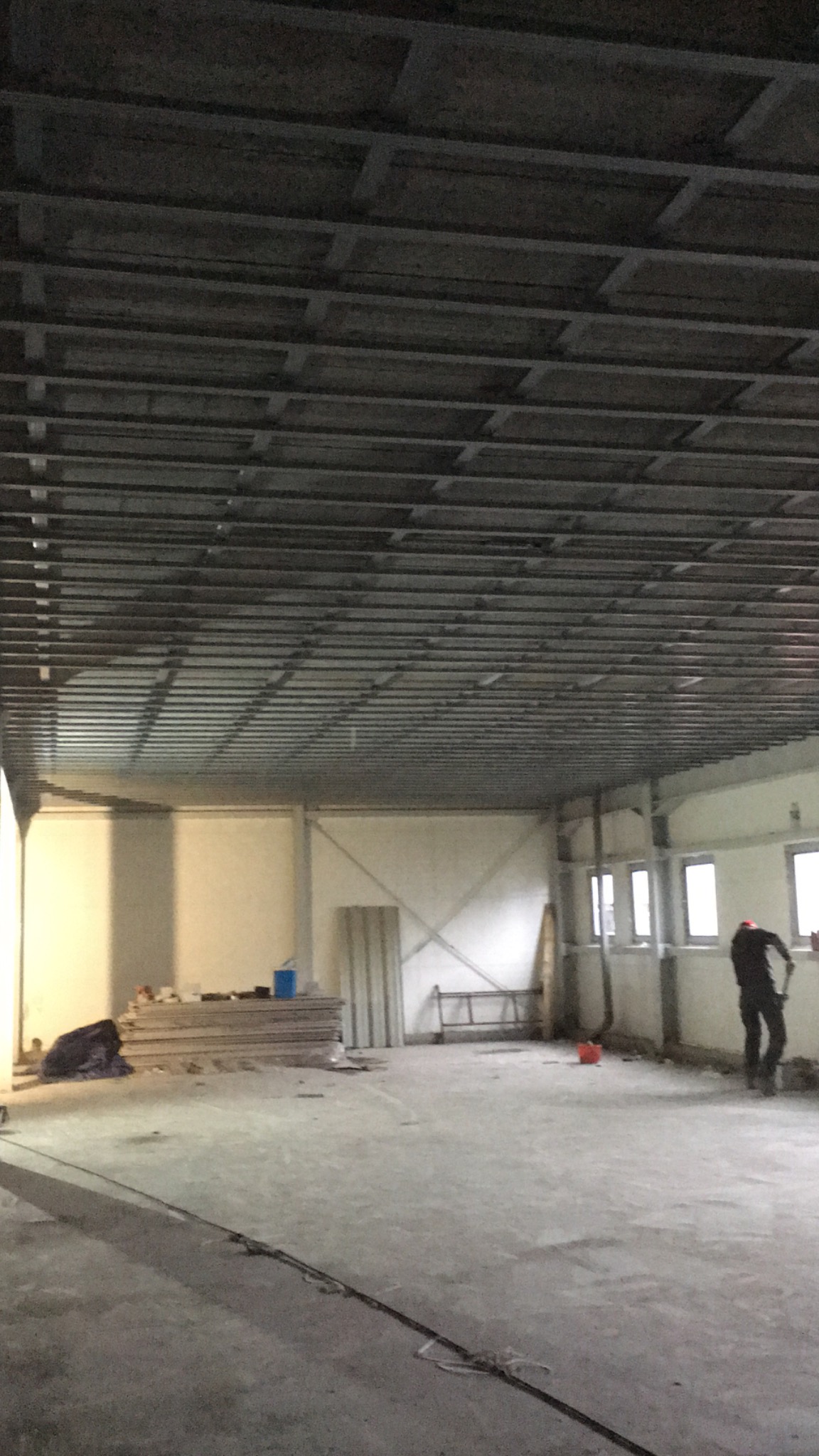 Duże, puste pomieszczenie w trakcie prac wykończeniowych, widoczna metalowa konstrukcja sufitu podwieszanego, stos płyt na podłodze, drabina i pracownik w oddali.