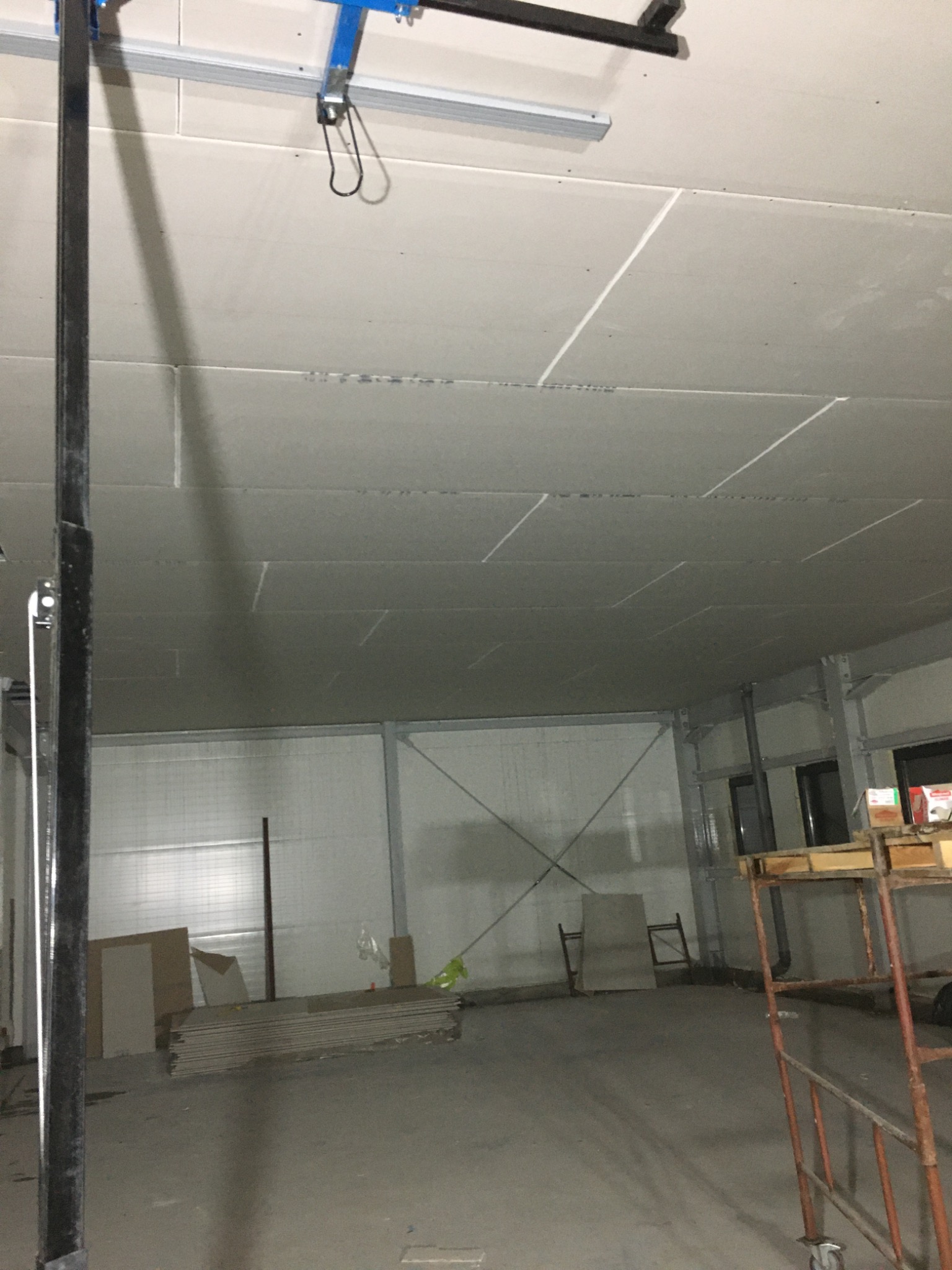 Montaż płyt kartonowo-gipsowych na suficie w dużym pomieszczeniu, widoczny podnośnik do płyt i stos płyt na podłodze.