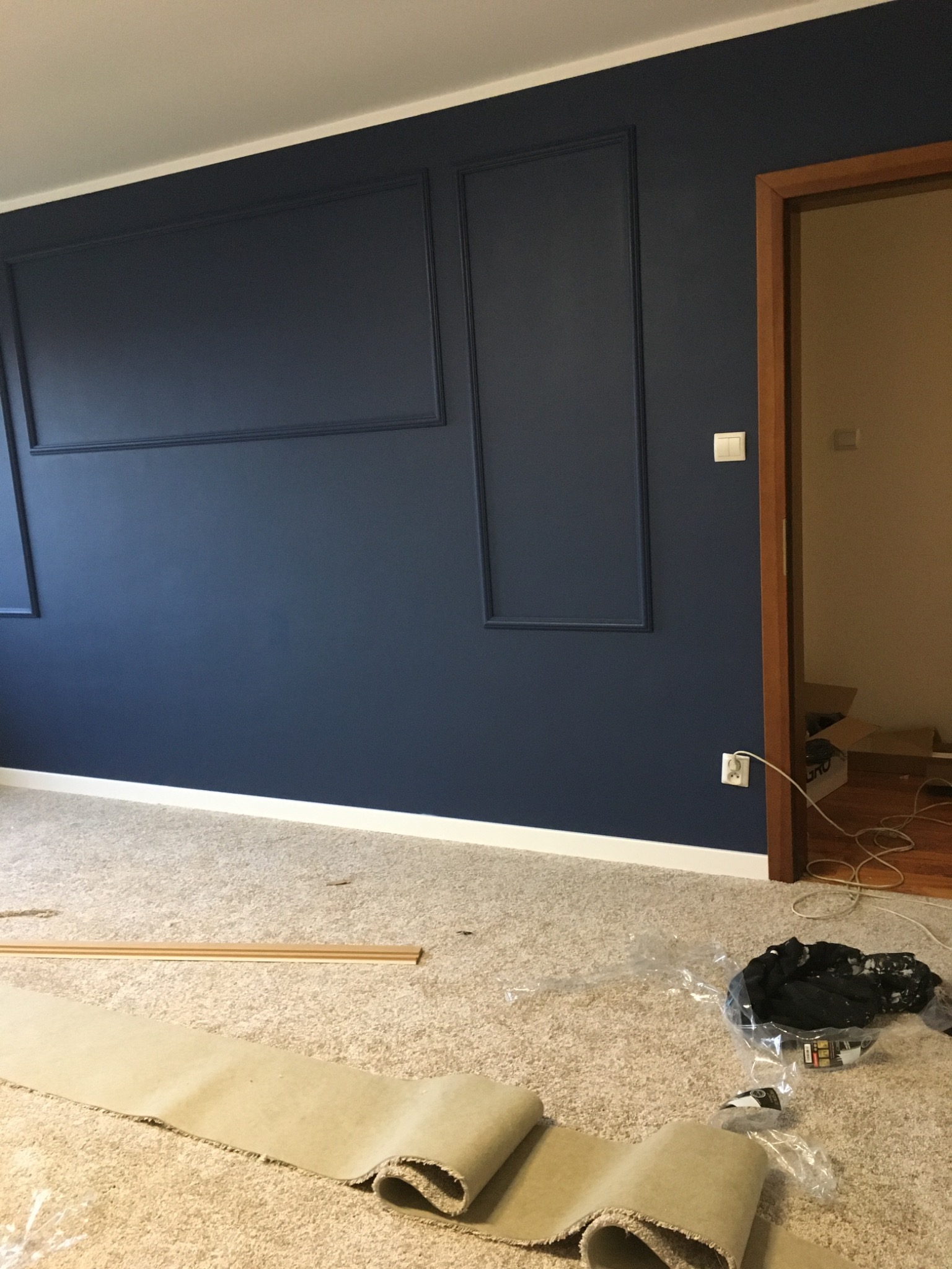 Niebieska ściana z dekoracyjnymi listwami w trakcie wykańczania, widoczny fragment rozłożonej wykładziny podłogowej i listwa przypodłogowa.