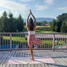 Kobieta w pozycji drzewa na macie do jogi na drewnianym tarasie z widokiem na zielony krajobraz z górami w tle.