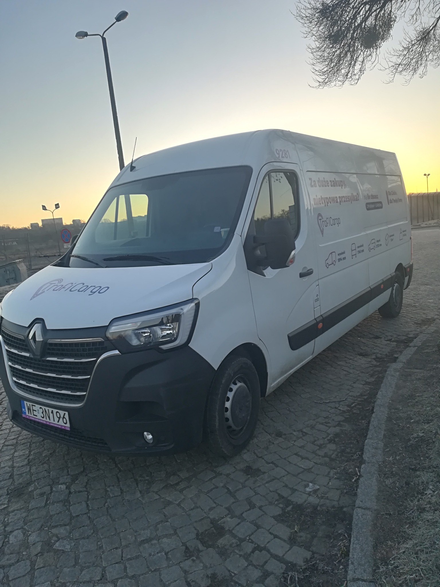 Biały bus Renault Master z logo firmy przewozowej ProfiCargo zaparkowany na brukowanej nawierzchni, widoczny numer rejestracyjny, sylwetki różnych rozmiarów pojazdów na boku, w tle latarnia...