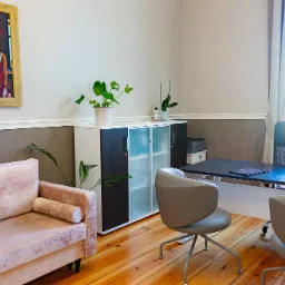 Jasne biuro z drewnianą podłogą, różową sofą, biurkiem z komputerem i obrazem w złotej ramie na ścianie.