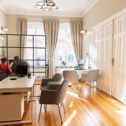 KELLER FINANSE - Jasne, przestronne biuro z drewnianą podłogą, białymi meblami i dużymi oknami z zasłonami; w tle widoczni dwaj mężczyźni za szklaną ścianką działową, siedzący przy biurku z komputerem.