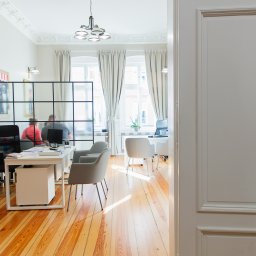 KELLER FINANSE - Jasne biuro z drewnianą podłogą, widok przez otwarte białe drzwi z ozdobną, brązową klamką, w tle biurka, krzesła i osoby przy komputerach.