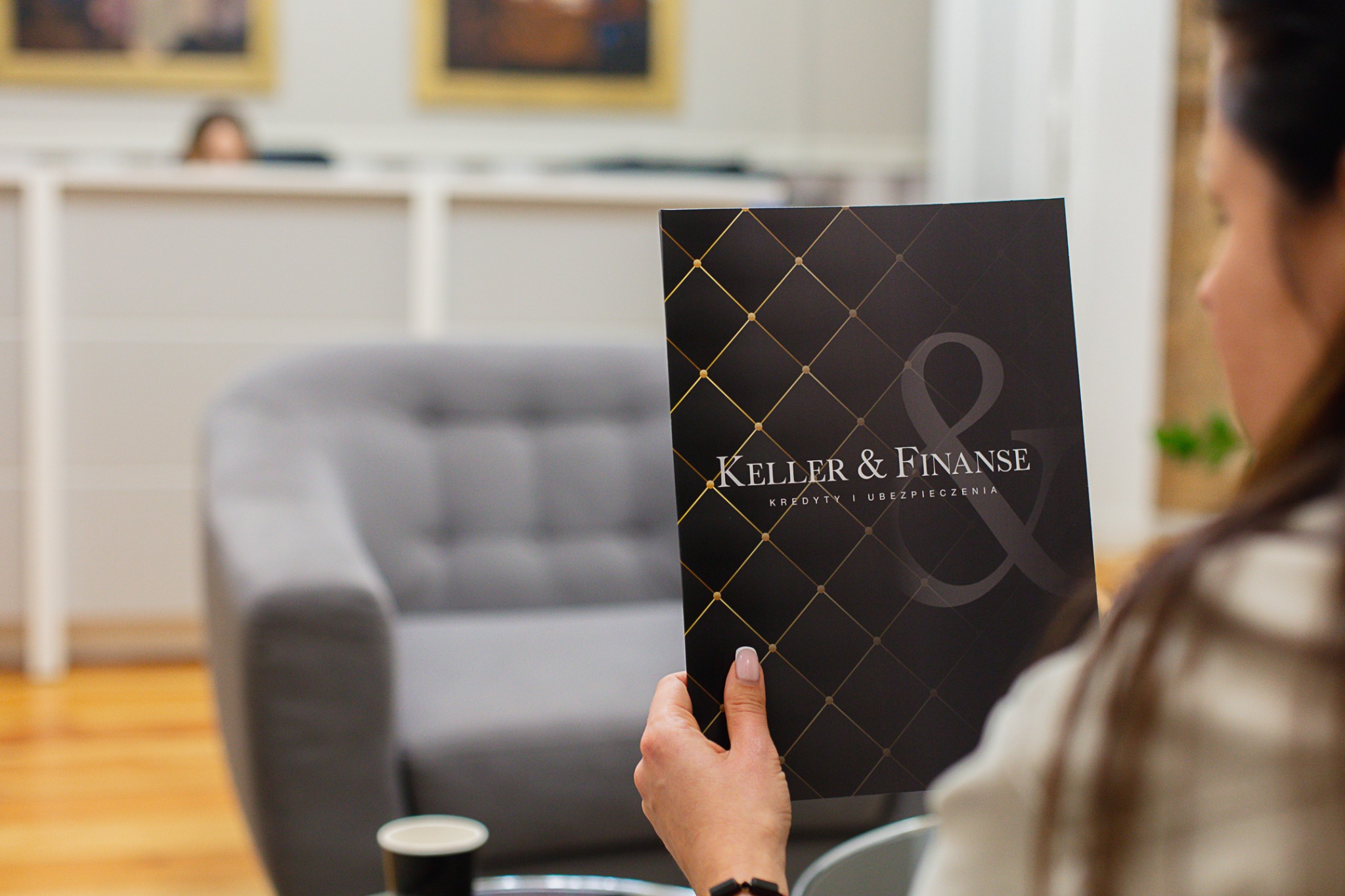 Kobieta przegląda czarny folder z logo 'Keller & Finanse' i napisem 'Kredyty i Ubezpieczenia' w nowoczesnym biurze z szarym fotelem.