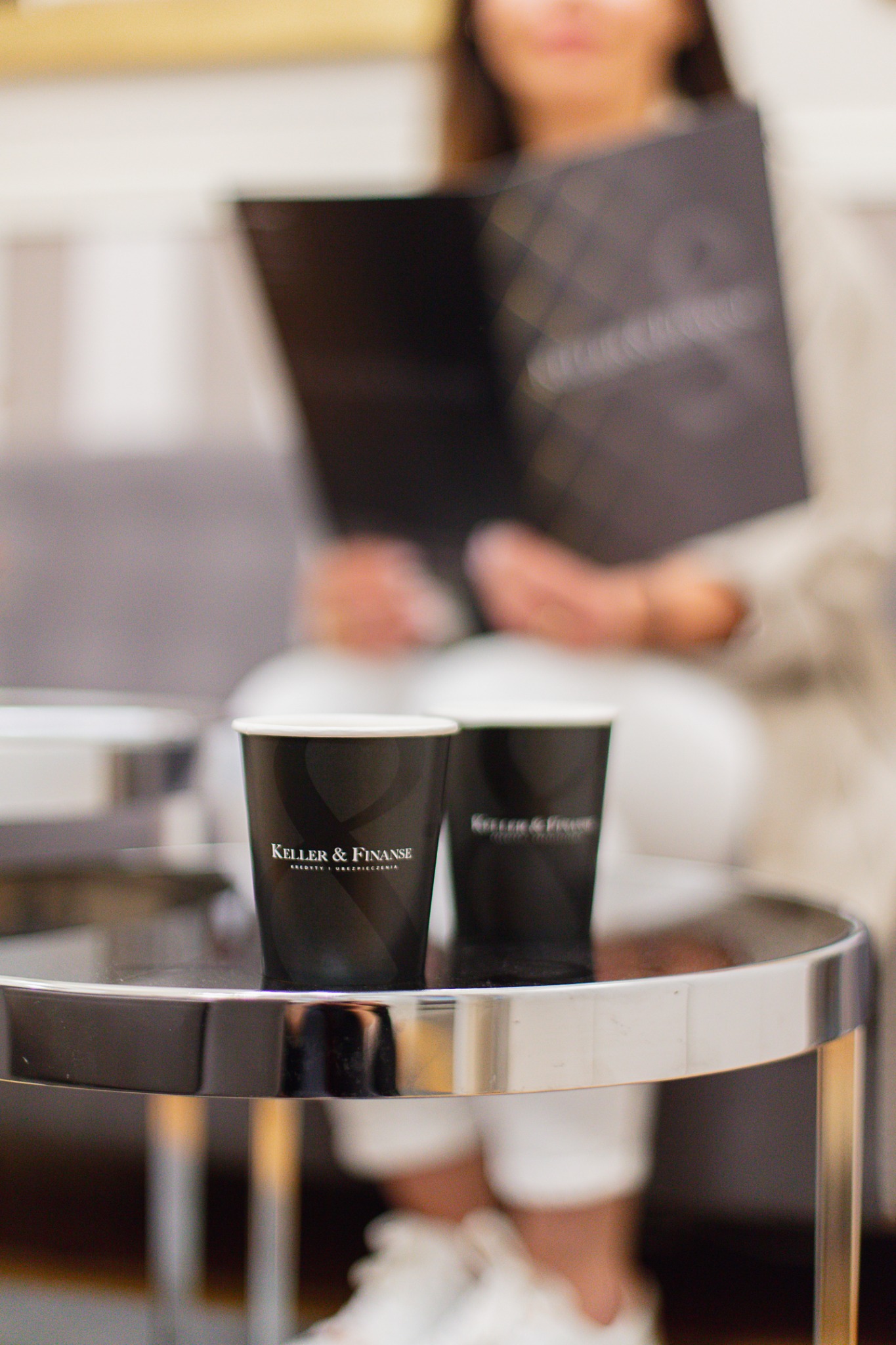 Dwa czarne kubki z logo 'Keller & Finanse' na okrągłym stoliku z metalowymi nogami, w tle kobieta przeglądająca dokumenty.