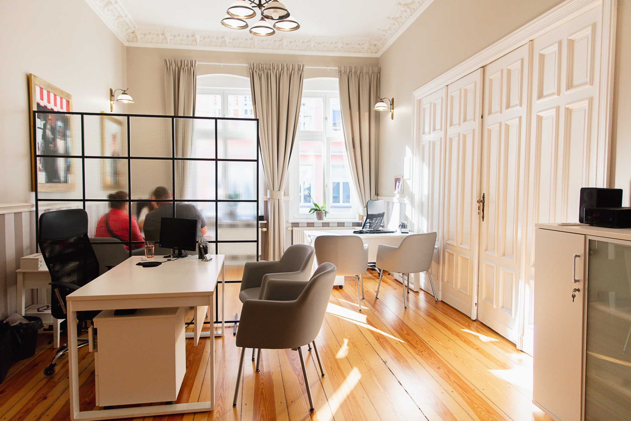 Jasne, przestronne biuro z drewnianą podłogą, białymi meblami i dużymi oknami z zasłonami; w tle widoczni dwaj mężczyźni za szklaną ścianką działową, siedzący przy biurku z komputerem.
