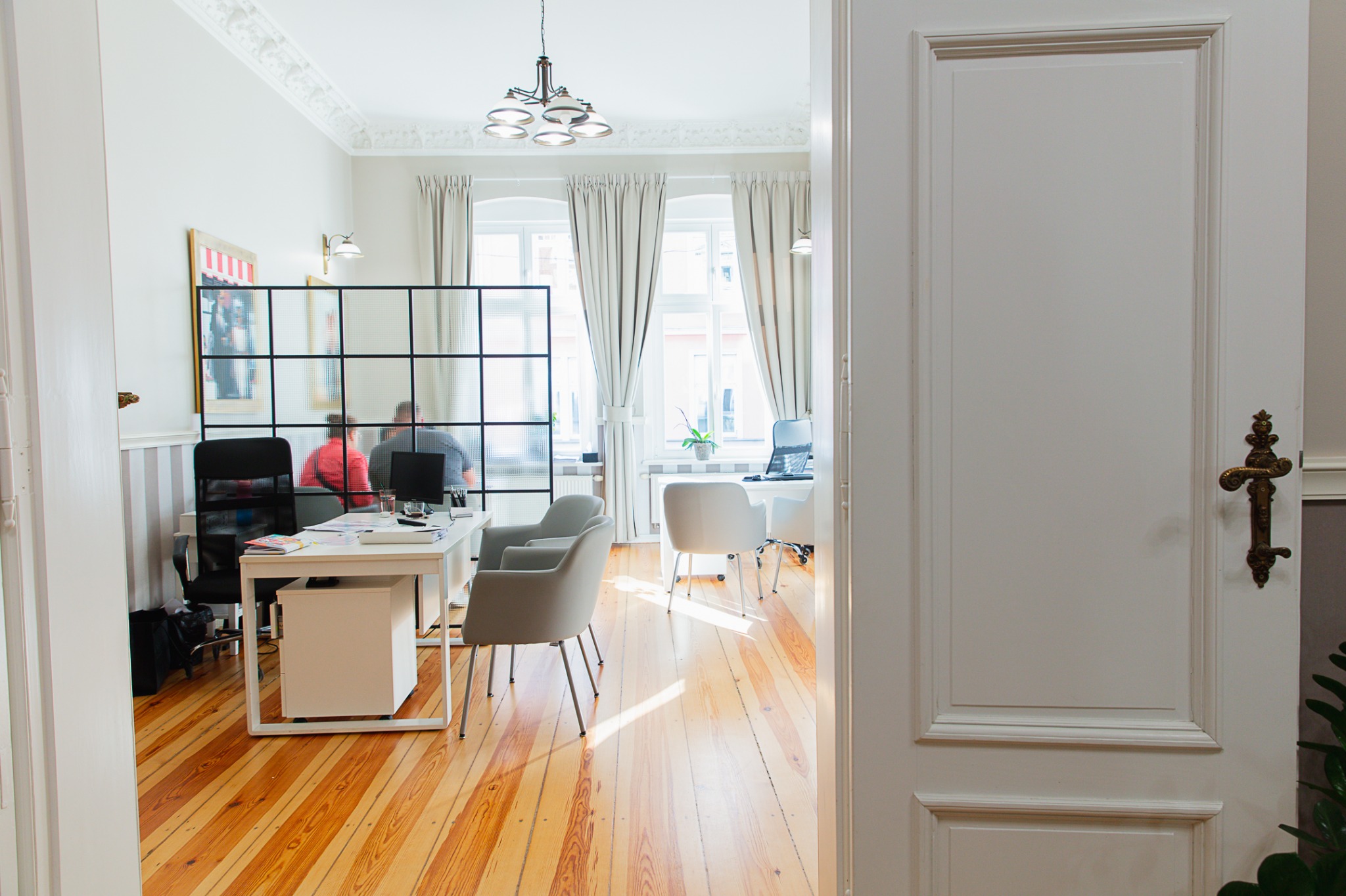 Jasne biuro z drewnianą podłogą, widok przez otwarte białe drzwi z ozdobną, brązową klamką, w tle biurka, krzesła i osoby przy komputerach.