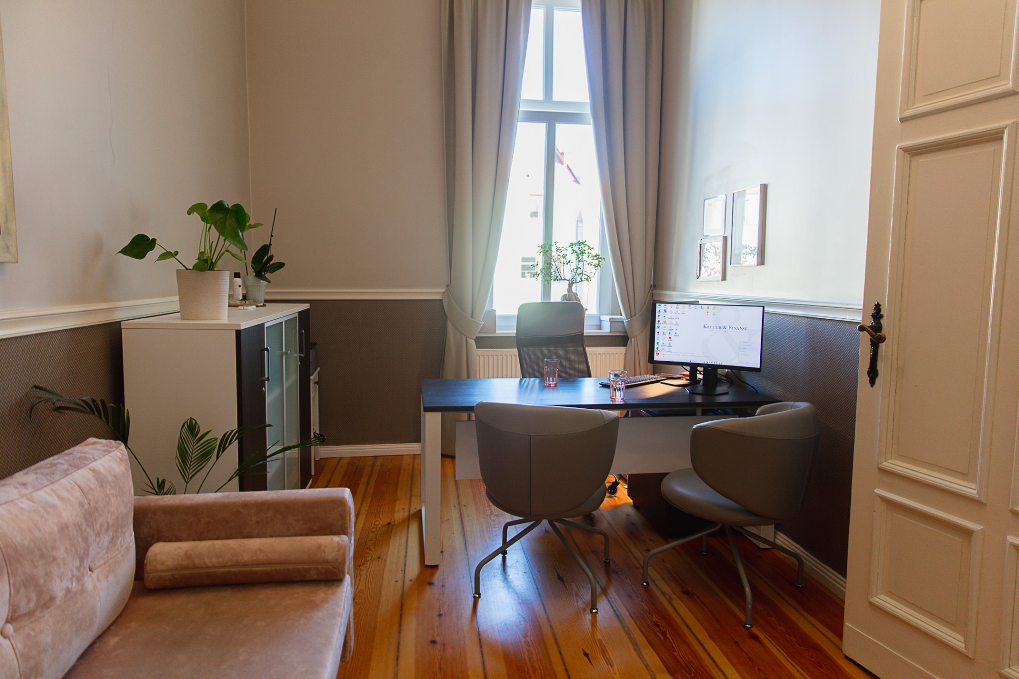 Biuro doradcy finansowego w Zielonej Górze z drewnianą podłogą, biurkiem, dwoma krzesłami, kanapą, szafką i roślinami doniczkowymi, widok na okno z firankami.