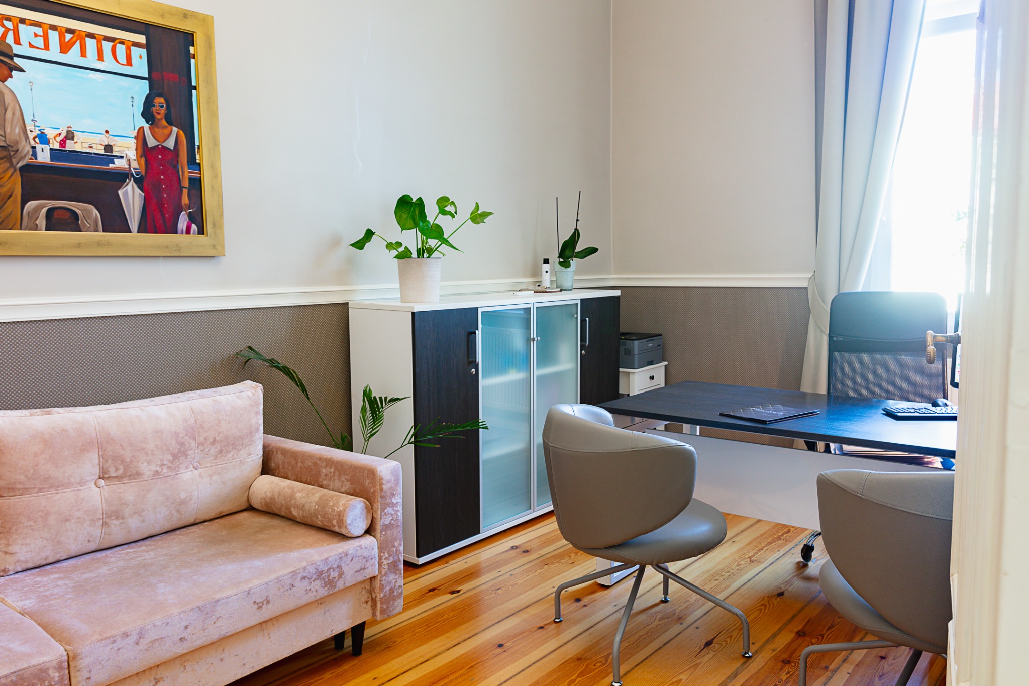 Jasne biuro z drewnianą podłogą, różową sofą, biurkiem z komputerem i krzesłami, szafką z roślinami oraz obrazem na ścianie.