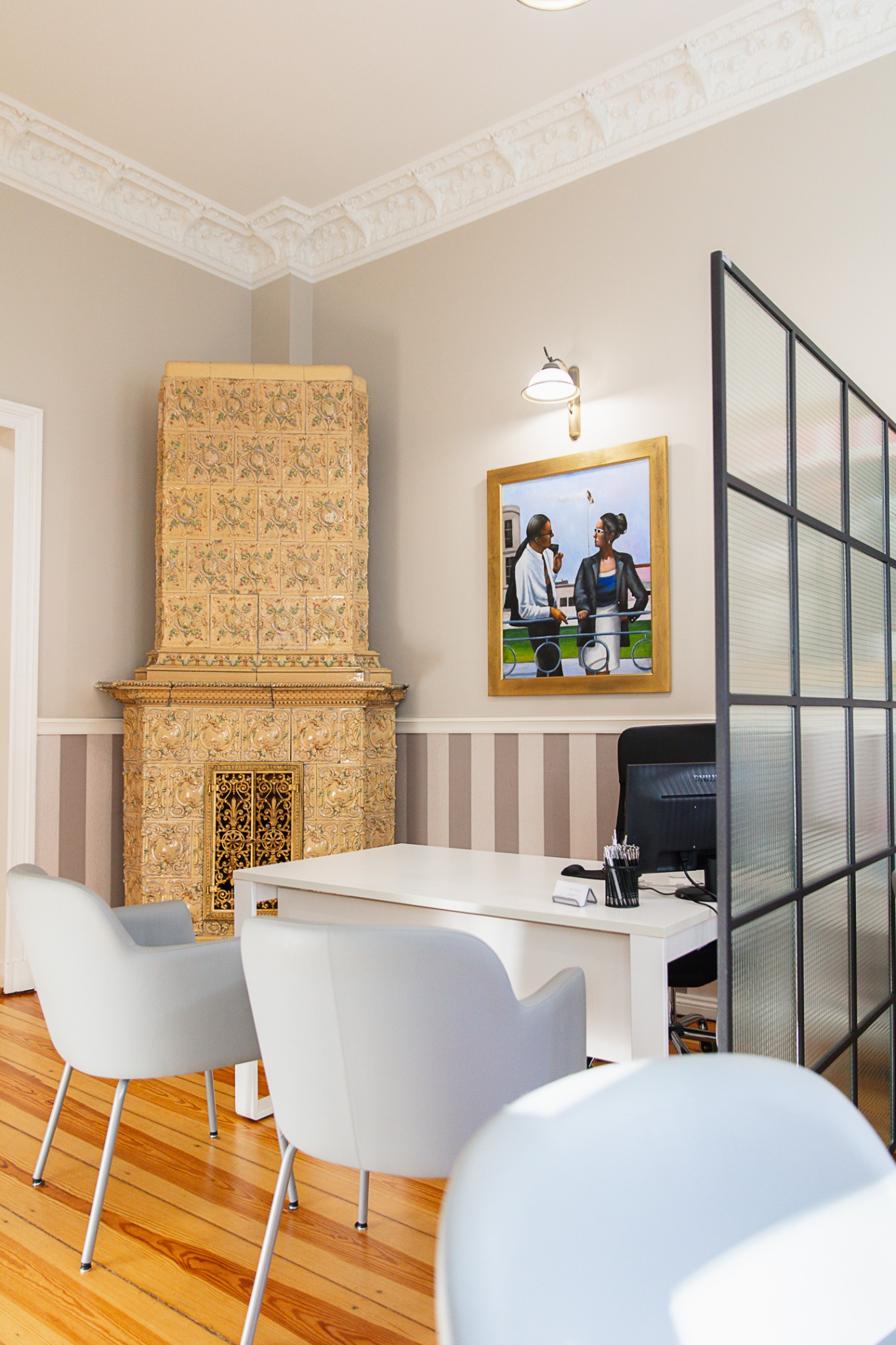 Jasne biuro z dużym, zdobionym piecem kaflowym, białym biurkiem, szarymi krzesłami i obrazem w złotej ramie przedstawiającym rozmowę biznesową.