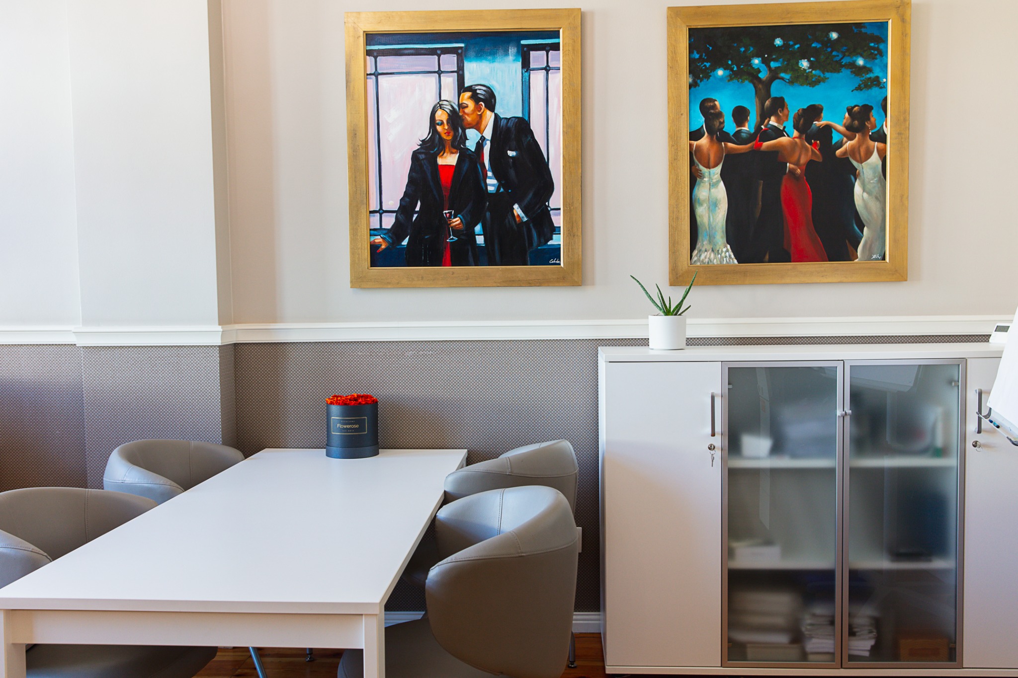 Jasne biuro z białym stołem, szarymi krzesłami, szafką z przeszklonymi drzwiami i dwoma obrazami w złotych ramach na ścianie.