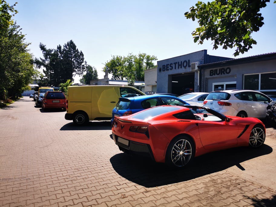 Czerwony sportowy kabriolet zaparkowany przed warsztatem samochodowym Besthol z innymi pojazdami w tle, widok z tyłu.