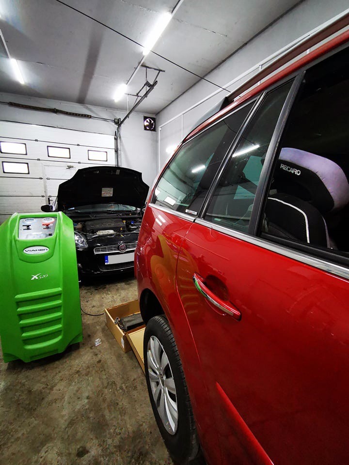 Wnętrze warsztatu samochodowego: czerwony samochód z fotelikiem Recaro, otwarty przód czarnego samochodu i zielone urządzenie Viahen X520 do obsługi klimatyzacji.